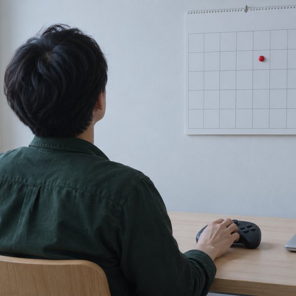 作業机でゲームコントローラーに手を置き、壁のマス目の予定表に付いた赤いマグネットを見る後ろ姿
